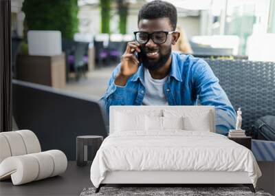 Portrait of happy african man using phone while working on laptop in a restaurant Wall mural